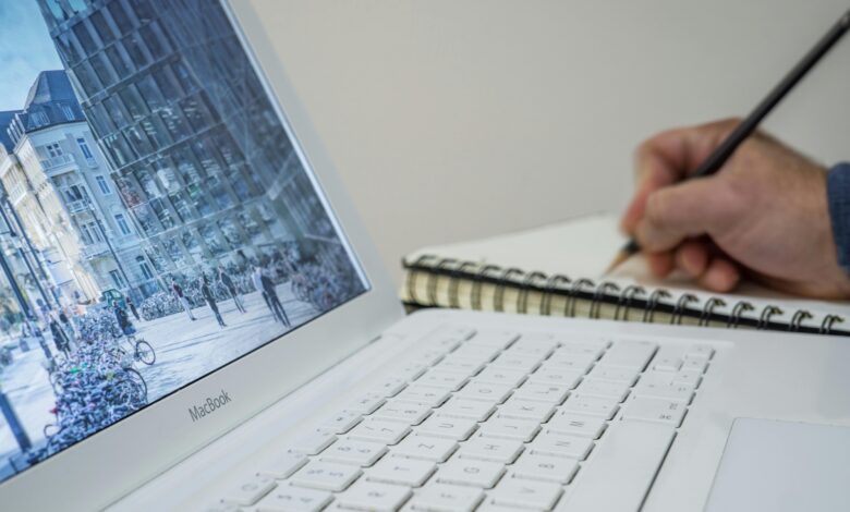 A person taking notes in a notebook beside an open laptop displaying an urban scene.