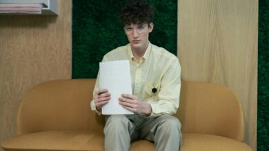 Young man sitting on a sofa in an office holding papers, wearing glasses and a yellow shirt.